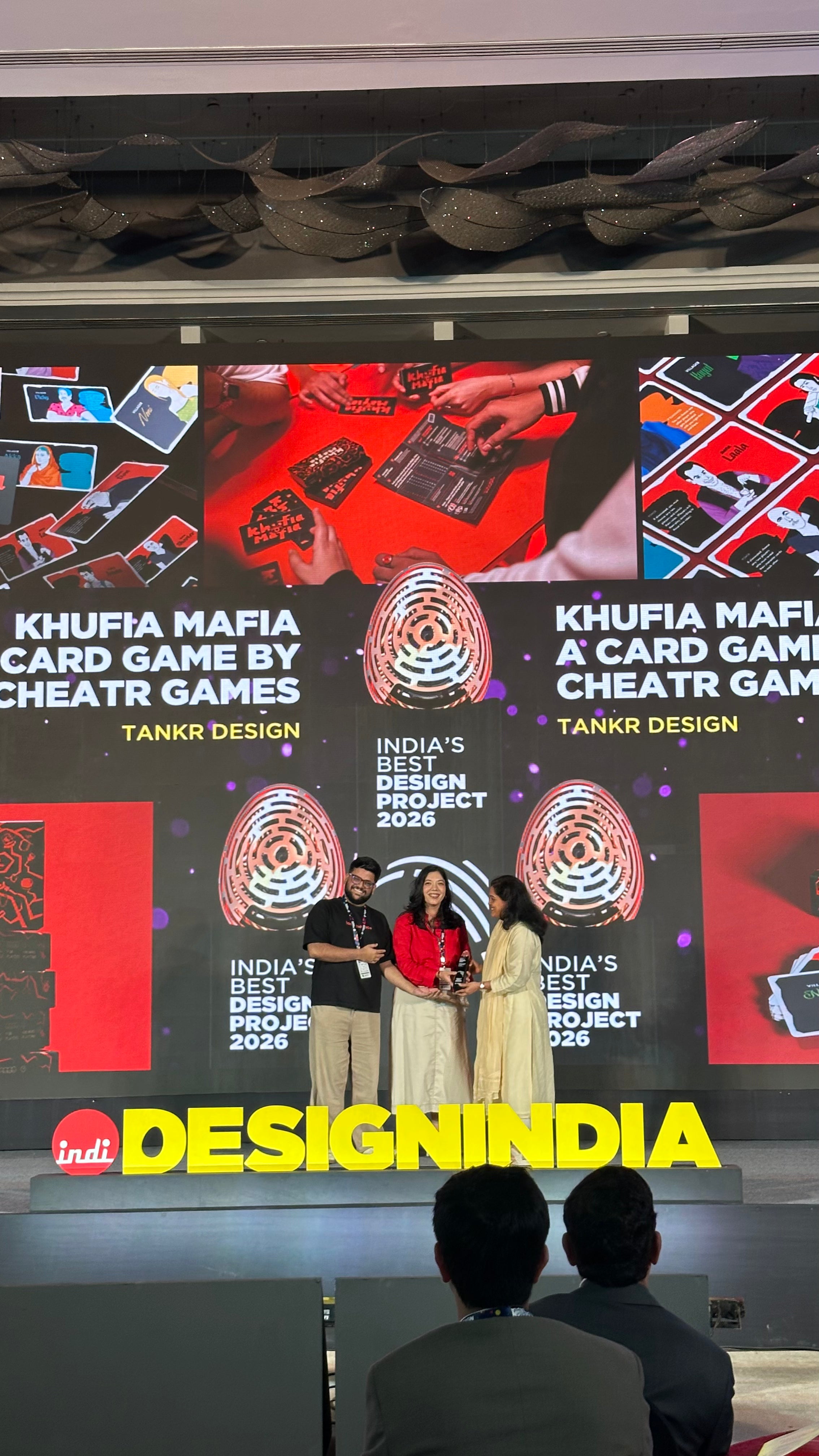A large event screen displays branding for 'Khufia Mafia Card Game by Cheatr Games' and 'India's Best Design Project 2024' by Tankr Design, with three people standing in the center under a red umbrella. The foreground shows the backs of audience members' heads, and a yellow 'DesignIndia' logo is visible at the bottom.